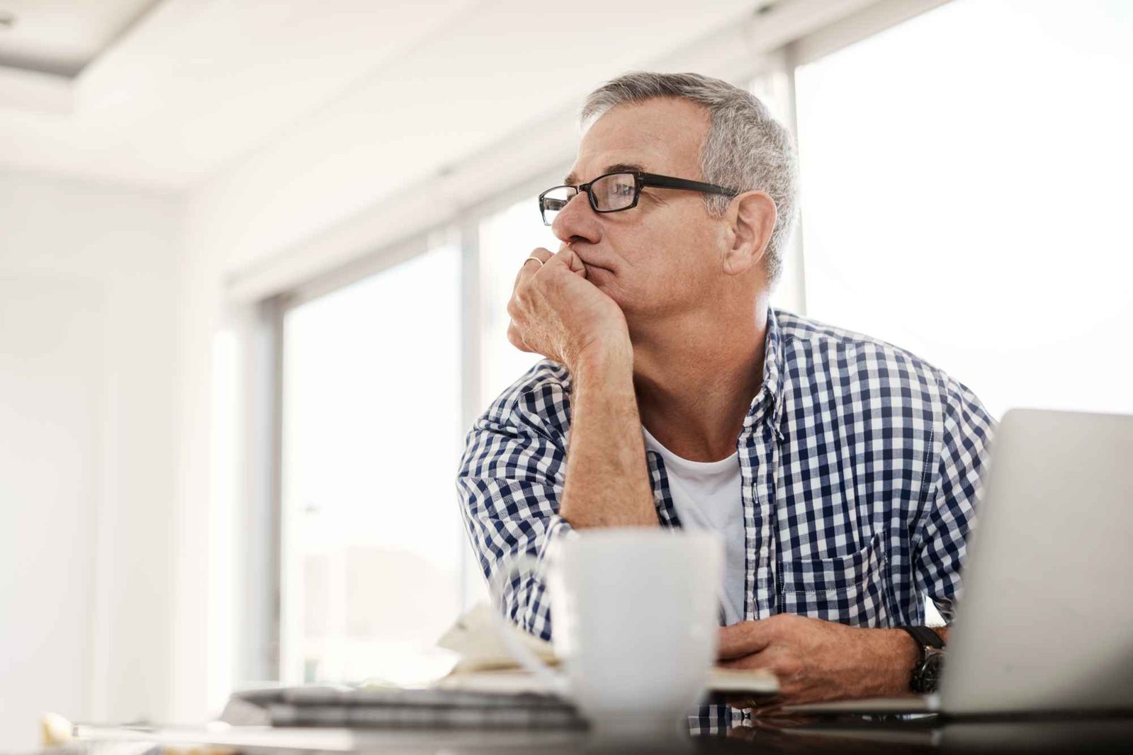 Mann mit Kaffeetasse am Laptop stützt Kopf in Hand und blickt nachdenklich zur Seite.